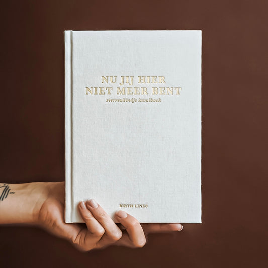Birth Lines sterrenkindje invulboek Nu jij hier niet meer bent met beige linnen kaft en goudfolie titel, vastgehouden in een hand tegen warme bruine achtergrond